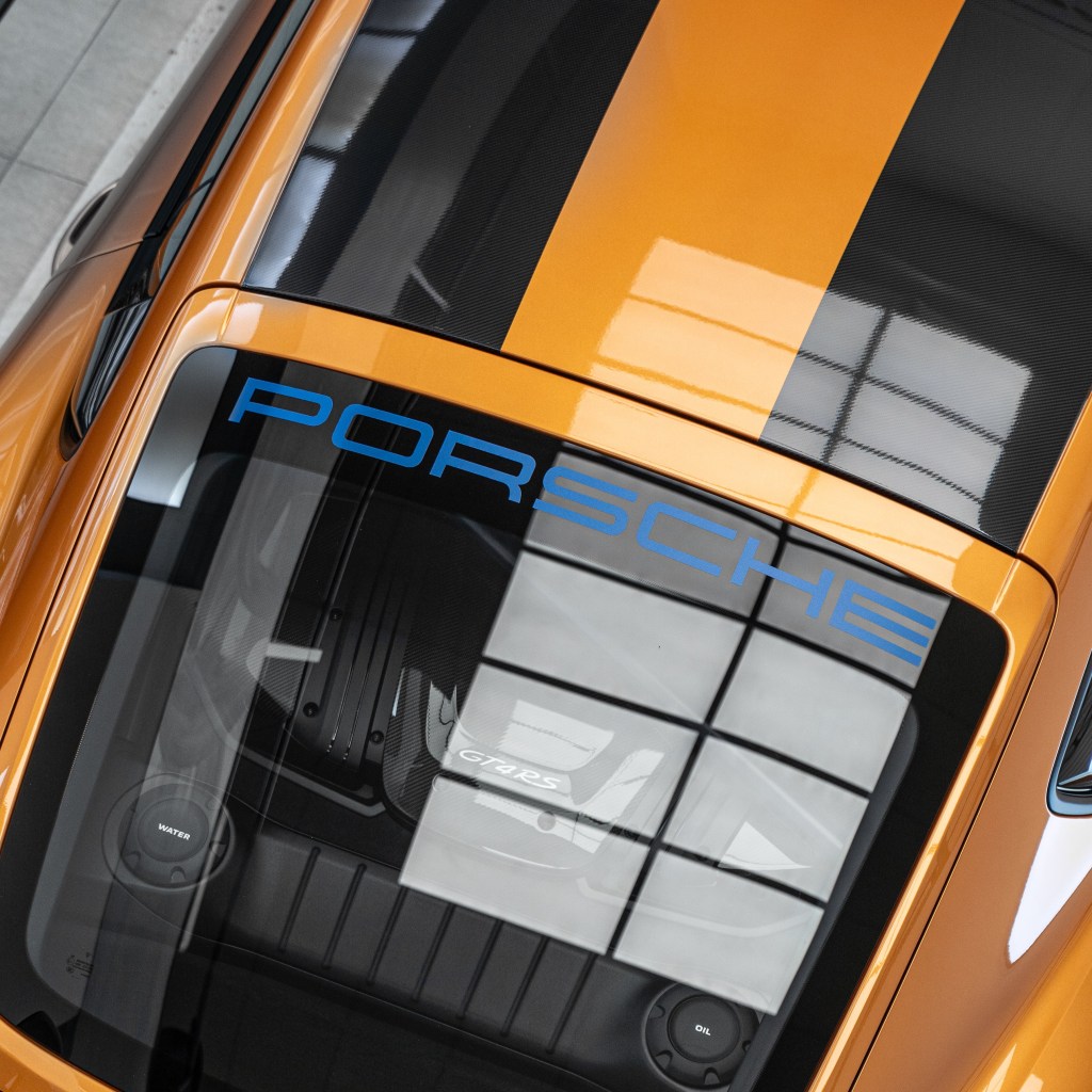 Top view of an orange Porsche car with a distinctive black stripe and the word 'PORSCHE' in blue on the rear glass, showcasing the engine and dashboard indicators.