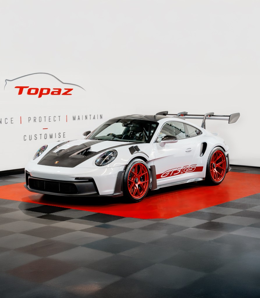 A white Porsche GT3 RS with a black roof and red wheels, displayed on a black and red checkered floor. The background features branding for Topaz, a car care service.